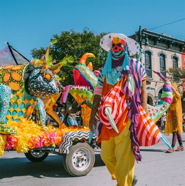 A person in costume standing on the street in front of a decorated vehicle.
