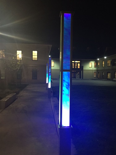 A long rectangular sculpture lit blue from within, installed along the walkway to an art studio entrance.
