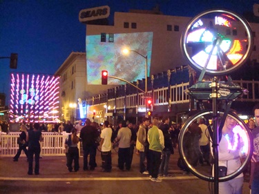 A number of art installations along a night street. People stand around the artwork. 