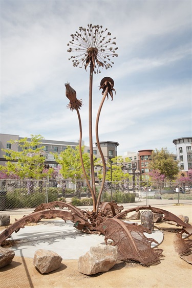 A metal sculpture of a dandelion with apartment buildings behind it. 