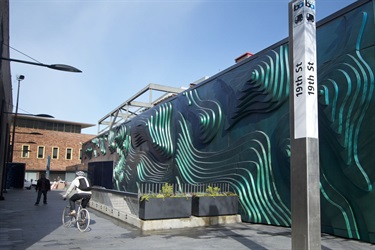 Dark green abstract sculpture on an exterior wall next to a BART train station entrance. 