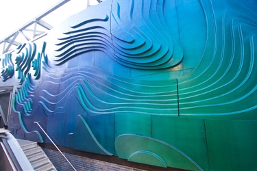 A green and blue abstract sculpture on an exterior wall next to a BART train station entrance. 