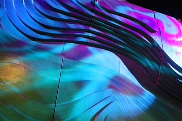 A green and blue abstract sculpture on an exterior wall next to a BART train station entrance. There is a purple light being projected on the sculpture.