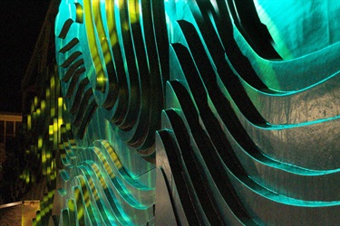 A green and blue abstract sculpture on an exterior wall next to a BART train station entrance. A yellow light is projected on the sculpture. 