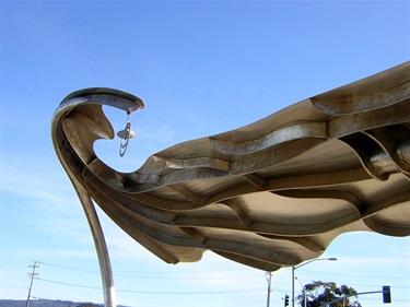 The top of a metal canopy sculpture in the shape of a wave, against a blue sky. 