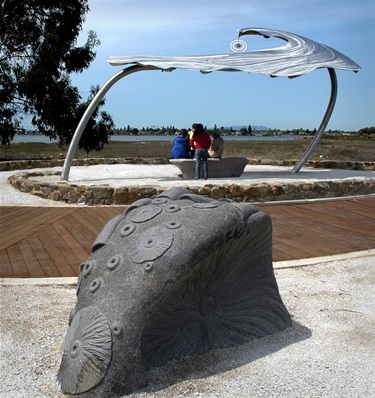 People gathered around a bench underneath a metal canopy sculpture. In the foreground is a stone sculpture with carved barnacles. 