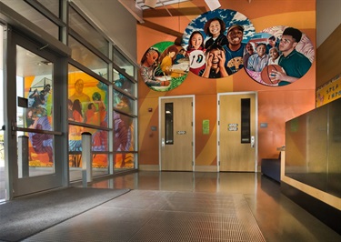 Large scale artwork showing groups of people, on medallions installed on the wall above the doors, inside of a recreation center lobby.