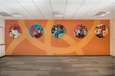 Five medallions with printed artwork, suspended on an orange wall inside a recreation center teen lounge. 